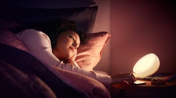 A woman lying in bed reaches over to the bedside table where there is a table lamp next to her phone