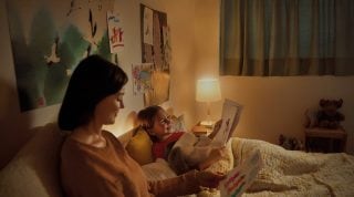 A young girl lies in bed while her mother sits on a chair beside her, the room is lit with dim light by a table lamp in the background